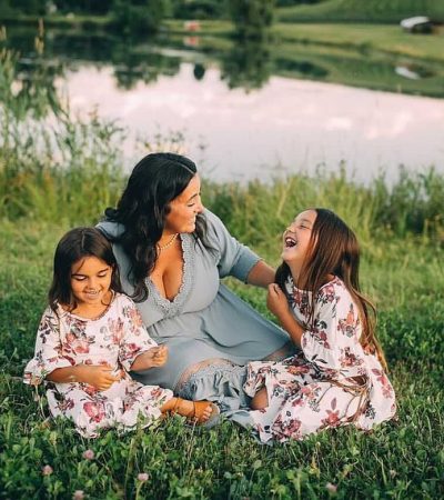 A woman and her two daughters sit in a field laughing with a pond in the distance